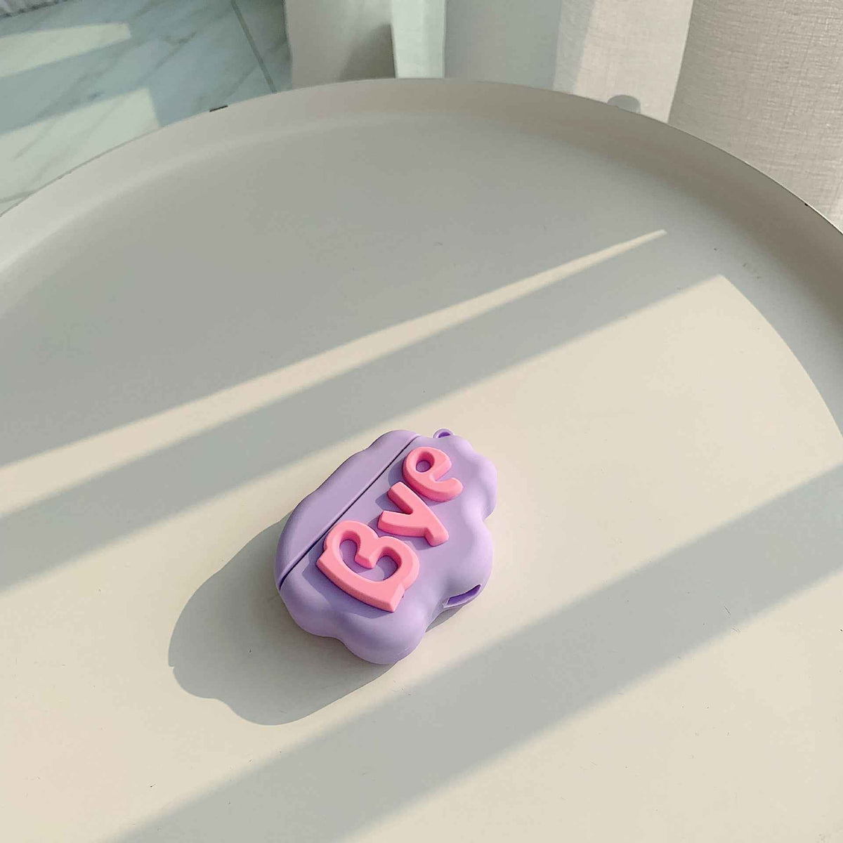 Purple silicone cloud AirPods case with raised “Bye” lettering, shown closed
