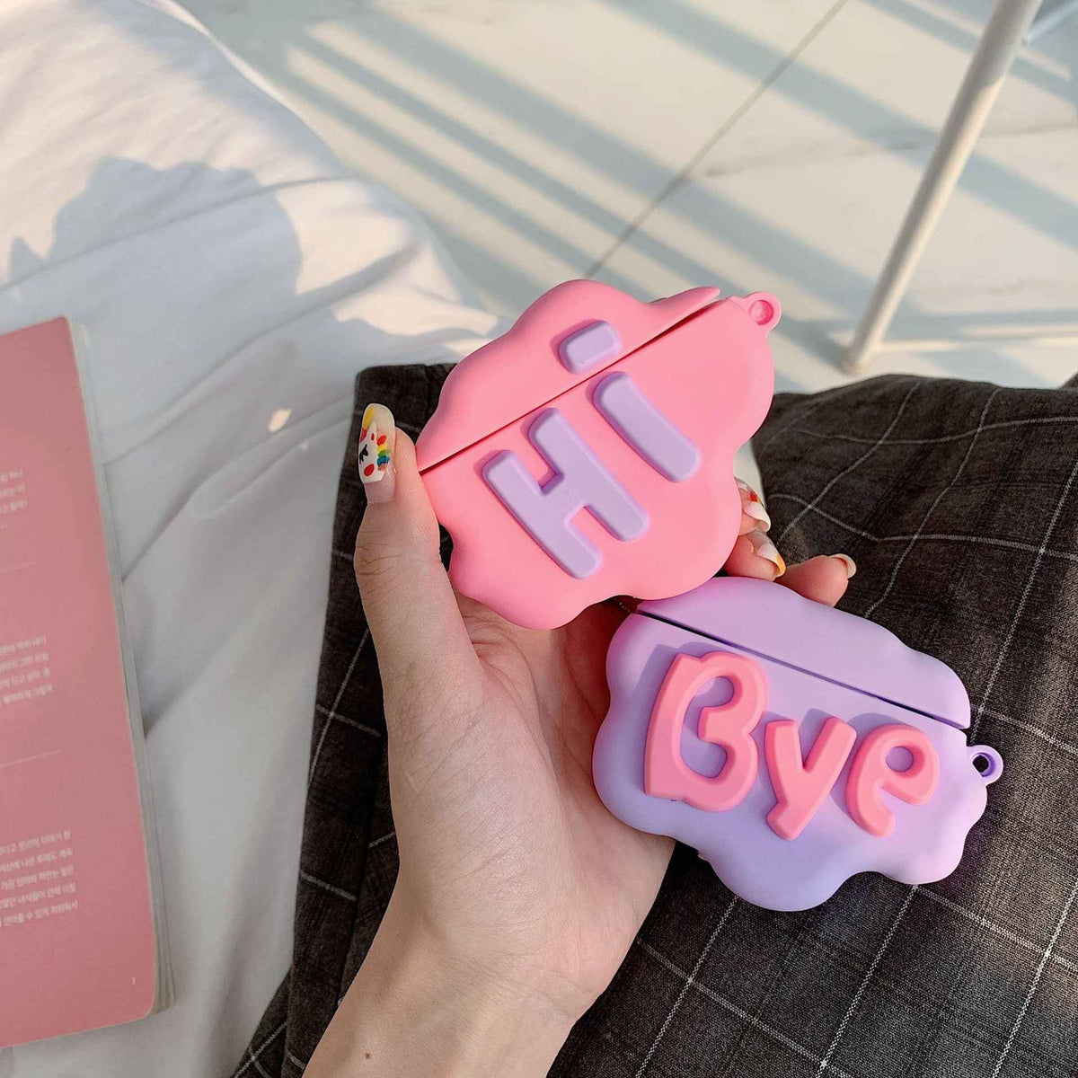Pink and purple silicone cloud AirPods cases with “Hi” and “Bye” lettering, open on lap