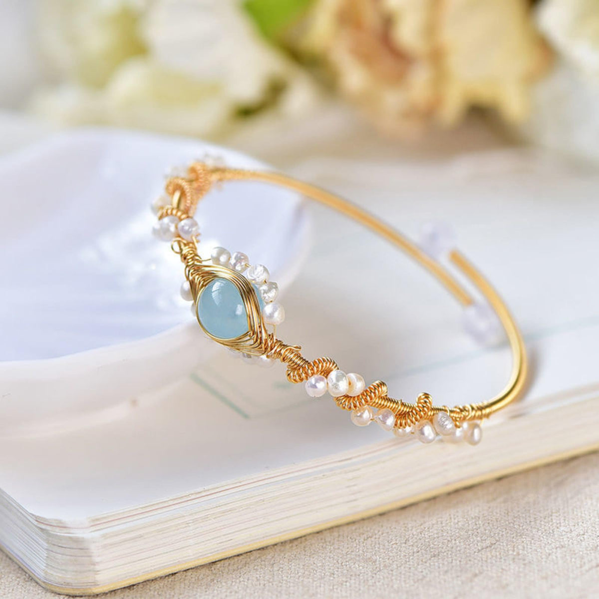 Gold wire cuff bracelet with white pearls, braided accents, and a blue oval charm