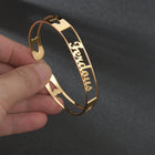 Gold stainless steel cuff bracelet held in hand; engraved cursive name on slim band